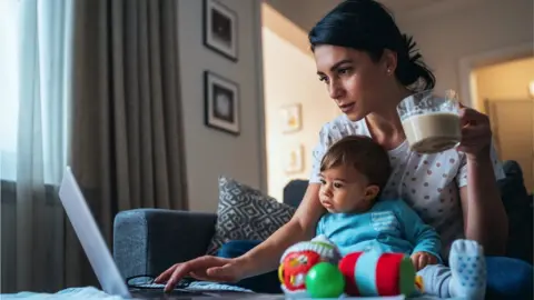 Getty Images A woman working from home with a baby sitting between her and her computer