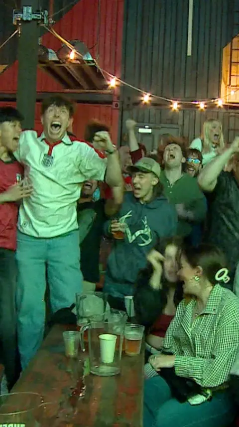 England fans celebrating as they watch the match. They are cheering and punching the air. One at the front is looking behind her at the jubilations.