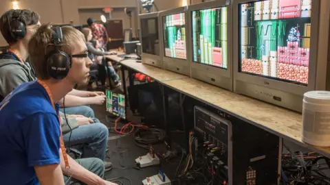 Games Done Quick Three people are playing games sat side-by-side. They are staring at their own separate monitors. On the floor are several SNES consoles connected to the TVs.