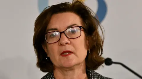 PA Media Eluned Morgan speaking at a microphone. she has dark rimmed glasses and hair, and is wearing a black jacket with white flecks in a fine pattern