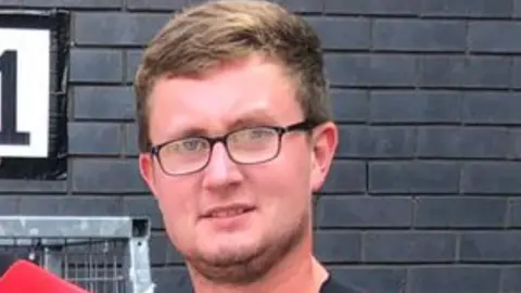 Jacob Hawden is photographed looking at the camera in front of a dark brick wall. He has short light brown hair and facial hair. He is wearing spectacles.