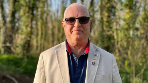 Joe Abbotts looking at the camera with trees behind him. He has short white hair, is wearing sunglasses, a cream blazer and a navy polo shirt.