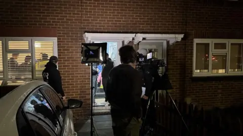 Bedfordshire Police A film crew outside a residential house at night. There is a lighting rig and a tripod with a camera, and people acting inside the hallway and kitchen. A man is standing next to the main camera and another man is lurks outside the front door.