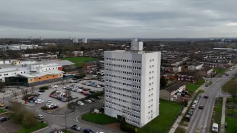 An aerial view shows a white tower block of around 10 storeys standing in a suburban setting under an overcast sky. The building is surrounded by a car park, low-rise housing and a community building with coloured cladding. Other tower blocks and rooftops are visible in the distance across the flat urban landscape.
