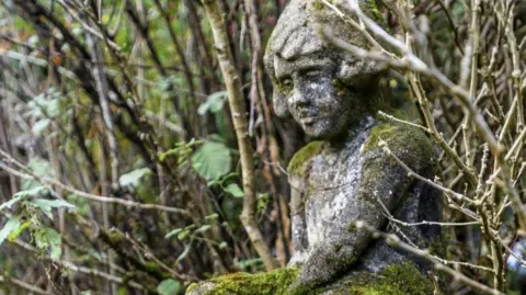 Ashdown Forest Foundation A small moss-covered stone statue of a seated child figure, partially hidden among branches and leaves in woodland at Cotchford Farm.