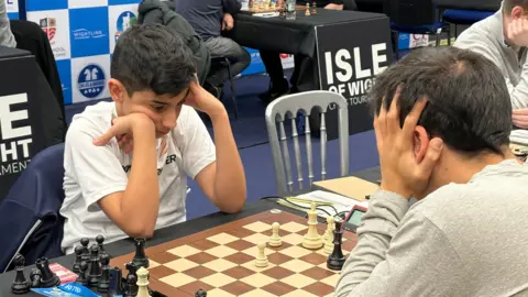 Rami, a young boy with dark hair, is playing a game of chess. He's leaning his head on his hands and staring intently at the board.