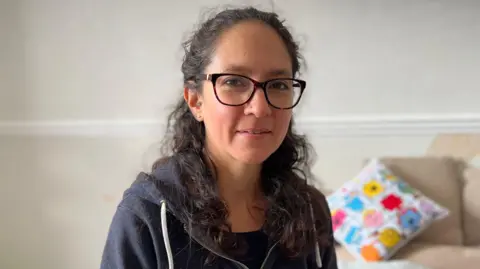 Ana Undah, who has  brown curly hair and glasses, is sat in her living room looking at the camera