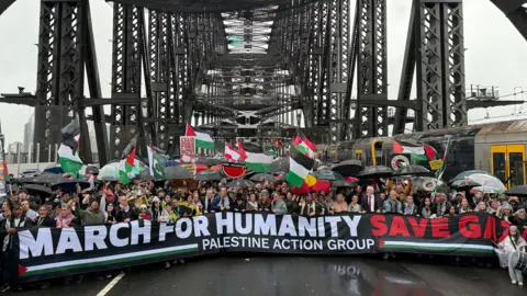 Dean Lewins/EPA centenas de manifestantes se reúnem sob os arcos da ponte do porto de Sydney, usando capas de chuva e segurando bandeiras vermelhas, verdes, pretas e brancas. Eles estão segurando um grande sinal que lê "Março para a humanidade: salvar Gaza".