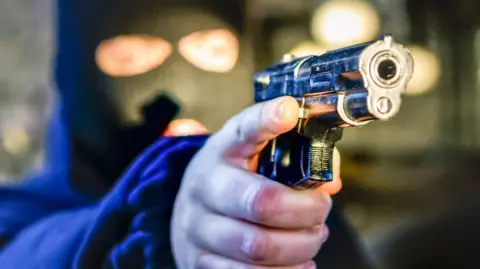 A man dressed with a balaclava over his head, in the style of a Northern Irish paramilitary, points a pistol handgun - stock photo