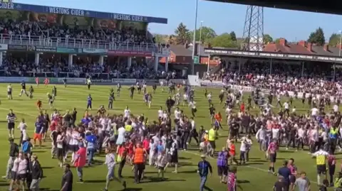 Fans of Hereford FC swarm onto the Edgar Street stadium football pitch. More than a hundred people appear to be on the pitch around the centre circle.