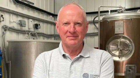 Alan Ridealgh, standing in a brewhouse, looking straight to camera. He is wearing a grey collared t-shirt. His arms are folded and he has short, grey hair. In the background are large metal containers, which are used in the brewing process. The walls are white corrugated metal.