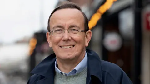 Martin Tod Man with glasses and short dark hair looking at the camera - he is wearing a navy coat, gre jumper and blue shirt.
