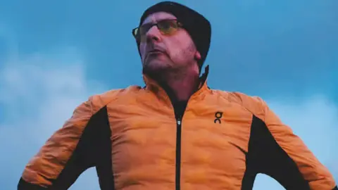 Head and shoulders of Ben Constable in running gear - an orange and black top, black hat and yellow lensed glasses against a blue sky