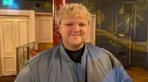 Daniel has short bleached blonde curly hair. He is smiling at the camera and is wearing a blue jacket over a black t-shirt.