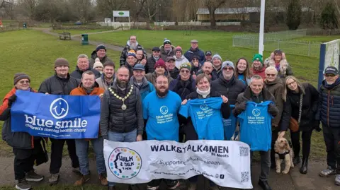 Walk & Talk 4 Men St Neots A group of people stood outside in a park. They are looking up at the camera and smiling. Those on the front row are holding blue signs or T-shirts with Blue Smile on the front, along with a blue smiley face. A white sign with Walk & Talk 4 Men St Neots is also being held at the front of the group. There are both men and women - and a dog - included in the crowd of people. 