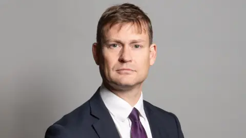 UK Parliament MP Justin Madders posing for an official parliamentary photo, wearing a navy suit and purple tie