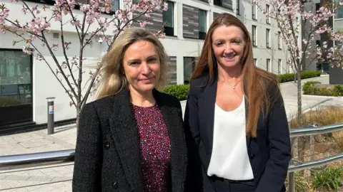 GIBA Helen Wyatt (L) and Jo Peacegood (R) are standing in a street smiling at the camera wearing formal jackets. There are two trees in pink blossom with grasses around them behind them and buildings beyond the trees.