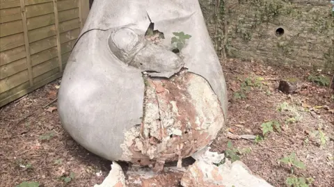 A stillborn and miscarriage statue in the garden, which is grey stone with a wrapped up baby in a blanket in the middle, with damage to the bottom of it. It is smashed.