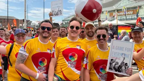 Three team members stand centre with others behind them. They wear yellow and red tops with Pride in Hull written on them. The person on the right holds up an A4 photo with "a game for everyone" written on it.