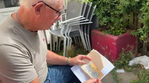 A man looks at a grainy yellow photo of of a truck. He is in a garden patio area with a red plant pot and grey chairs