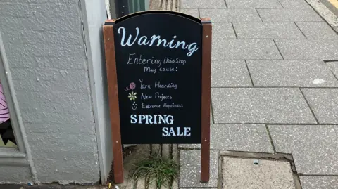 A sign outside a shop reads 'Warning: Entering this shop may cause yarn hoarding, new projects, extreme happiness. Spring Sale'