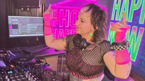 DJ Amyfist from the Brighton-based BBG Collective, pictured at her decks, headphones round her neck, wearing pink fluorescent fingerless gloves and black fishnet top - against a backdrop of neon pink and green signs.