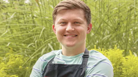 Josh Smalley, a young man with short fair hair. He is wearing a grey T-shirt under a black apron. He is looking at the camera and smiling.