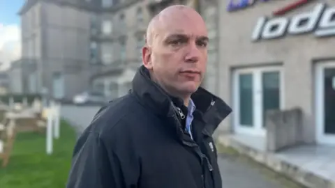 Louis Gardner looks into the camera wearing a black waterproof jacket and blue shirt 