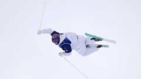 Mateo Jeannesson in action in the Freestyle Skiing Men's Moguls Qualification. He is horizontal in the air, holding two ski poles, with skis on his feet, and wearing white trousers, white top, with a helmet, goggles, and a blue vest. He is surrounded by white snow. 