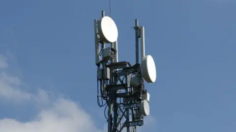 Mobile phone mast at back of farm yard in Carlisle, Cumbria. Only the top of the mast, which contains light coloured discs like satellite dishes, can be seen against a blue sky.