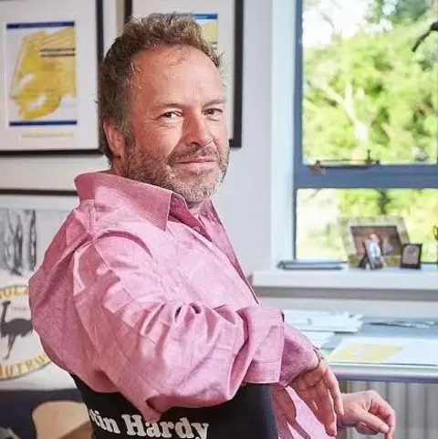 Hardy Pictures Justin Hardy, in a pink open-necked shirt, sits at a desk in front of a window. He is smiling over his shoulder and sitting in a director's chair with his name on it.