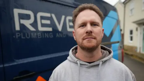 John Fairhall/BBC Ash Revett smiles at the camera in front of a blue van. He has short red hair and a red beard. He is wearing a grey hoodie. 
