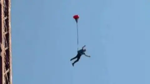A man jumping from Blackpol Tower. He is seen suspended in mid-air with a red balloon attached to him. Blue sky and the edge of the tower can be seen in the background.