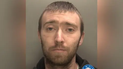 A mugshot of a man with short, brown hair and a beard. He is staring into the camera and wearing a black hoodie. 