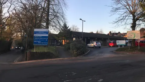 Google The exterior of Haslemere Community Hospital, showing the entrance sign, the driveway and two cars, one grey and one red, parked outside the building.