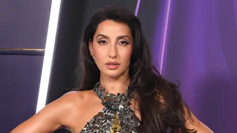 Nora Fatehi faces the camera and wears a grey sparkling sequined halter neck top. Her long brown hair falls on her shoulders and she stands against a purple background