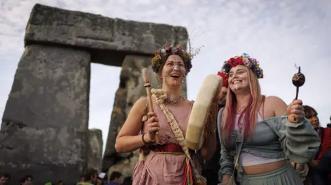 EPA Two women wearing flower crowns smile as one beats a handheld drum and another waves a shaker. Behind them is an arch formed by the stones of Stonehenge. 