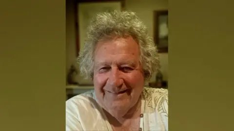 Andrew St. Helier Bourke Andrew St. Helier Bourke has grey hair and is smiling at the camera. He is wearing a white top with a green pattern on it. The background is blurred. There are framed pictures on the wall behind him.