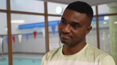 A man with cropped black hair and a trimmed beard stands on a viewing platform above a swimming pool. He is wearing a T-shirt with light green and white stripes.