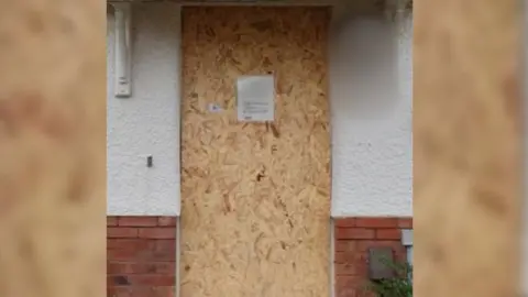 A boarded up door with an A4 printed sign on it which is too small to be read. A plywood board covers the door full. Beside it are white rendered walls with red bricks below.