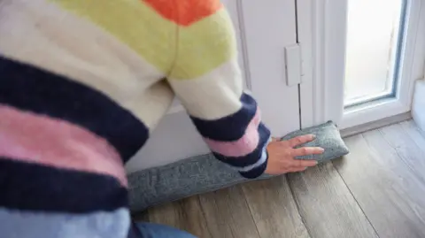 Getty Images A woman wearing a multi-coloured stripey jumper kneels in front of her front door as she fits a long grey draught excluder. 