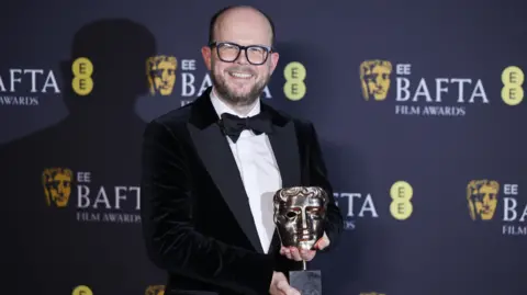 EPA Nick Emerson, who is bald and wearing black-rimmed glasses and a tuxedo with black bow tie and velvet black jacket, smiles while holding his Bafta award. Behind him is a screen with the Bafta logo.