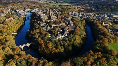 Aerial view of Durham in sunshine. The cathedral, castle and associated buildings stand in the bend of the river with the city spread out on either side behind. The scene is saturated in colour, with the river a deep blue, fringed by trees in a variety of autumnal colours.