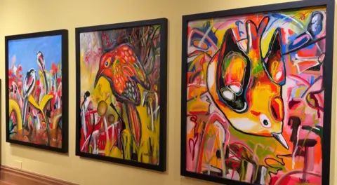 Michael Broomhead/LDRS Three vivid paintings depicting birds hang on a gallery wall.