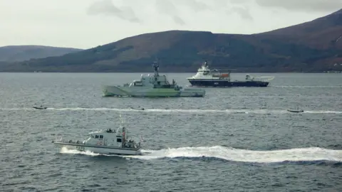 LPhot Daniel Bladen Three of the Rattlers can be seen moving through the water. They are surrounded by three larger vessels guiding them. In the distance is dark green and brown hillside.