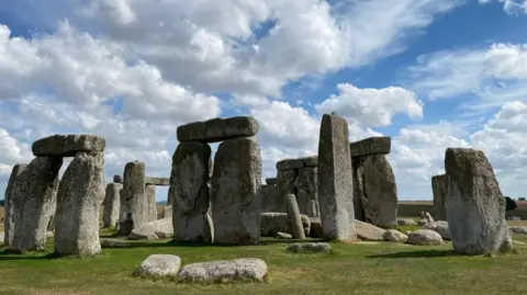 The famous hanging stones of Stonehenge against a bright blue sky dotted with clouds. 