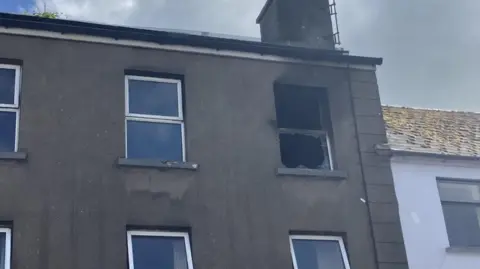 A grey building on Main Street in Ballymoney, with a chimney on top. The window on the far right is broken and there is evidence of smoke damage.