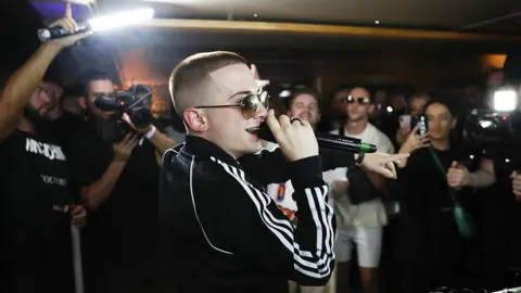 Getty Images ArrDee holding a balck microphone to his lips, while there are multiple people in the back holding camera phones, lights and cameras recording him. His left hand is pointing at the crowd. He is wearing dark sunglasses, with the side of his visible. He is also wearing black adidas tracksuit top with the three white stripes down the side of his right arm.