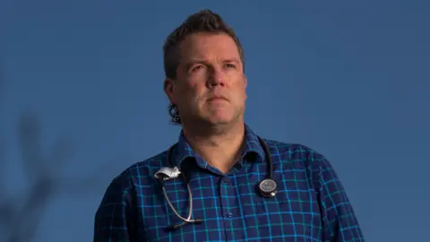Dr Chris Webster standing outside, a bare tree in the foreground. A stethoscope is around his neck and he is wearing a blue check shirt.