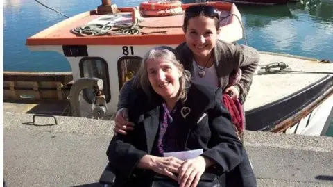 Pippa Marshall A smiling woman with long hair and wearing in a black coat sits in a wheelchair.
Her daughter stands behind her holding the chair. She has sunglasses pushed up on her head.
They are in front of a white boat with the number 81 on it.